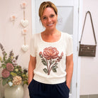 A smiling woman wearing a cream short-sleeve knit top with a large embroidered pink and green rose on the front, paired with navy pants, standing in a decorated indoor setting.