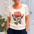 A woman wearing a cream-colored short-sleeve knit top with a scalloped scoop neckline and a large embroidered red and green rose on the front, paired with navy blue bottoms.