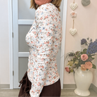 A woman wearing a fitted long-sleeve top with a small multicolor floral print on a white background, standing in an indoor room with a door, wall decorations, and a vase of flowers.