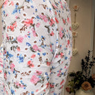 A close-up of a white textured fabric, likely a lace or embroidered tablecloth, featuring a colorful floral pattern with pink, blue, and red blossoms. The background shows a beige wall with decorative hanging hearts and a vase of dried flowers.