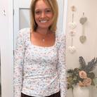 A smiling woman wearing a white long-sleeve scoop-neck top with a small pink, blue, and green floral print, paired with a dark brown skirt, standing in an indoor setting.