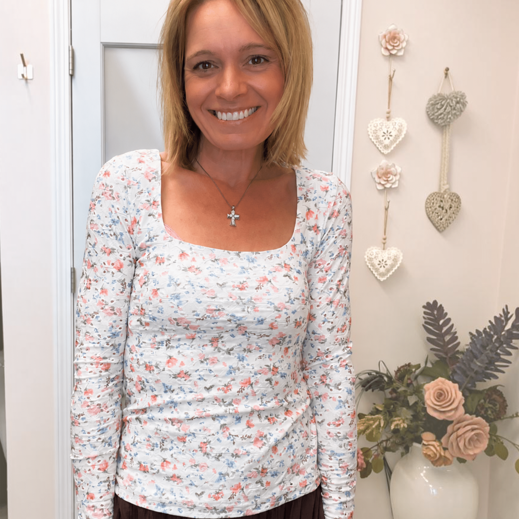 A smiling woman wearing a white long-sleeve scoop-neck top with a small pink, blue, and green floral print, paired with a dark brown skirt, standing in an indoor setting.