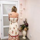 A woman stands with her back to the camera wearing a cream-colored maxi dress with a vibrant red, pink, and green floral print. She is positioned in front of a white door with a 'Dressing' sign, in a room with a light pink wall decorated with hanging flowers and a vase of dried flowers.