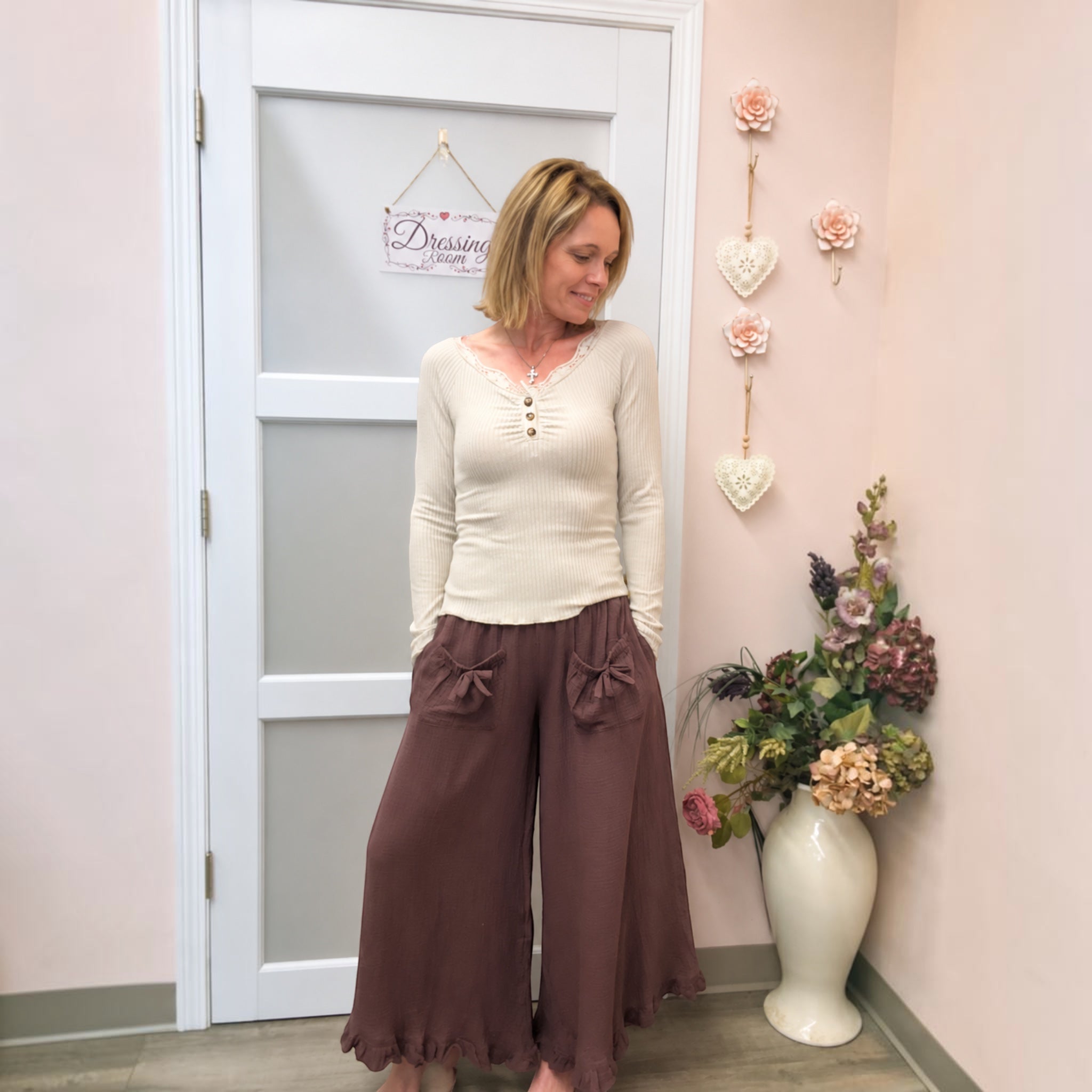 A woman stands indoors wearing a cream long-sleeve top with a buttoned henley neckline and dark brown wide-leg pants with bow details on the front. She is posed in front of a white door with a 'Dressin' Room' sign, against a light pink wall decorated with hanging flowers and a vase of faux flowers.