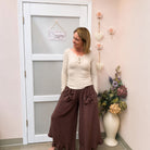 A woman stands indoors wearing a cream ribbed long-sleeve top with a buttoned neckline and dark brown wide-leg pants with bow details. She is posed in front of a white door in a pink-walled room with decorative wall flowers and a vase of artificial flowers.