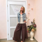 A woman modeling a casual outfit consisting of a light-wash denim vest over a cream ribbed top and brown wide-leg pants with bow details, standing in a room with a pink wall, white door, and a vase of flowers.