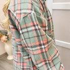 A close-up of a person wearing a long-sleeve plaid flannel shirt in sage green, pink, black, and white. The background shows a neutral indoor room with a vase of flowers.