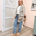A woman modeling a light green and pink plaid button-up shirt over a white t-shirt, paired with blue jeans and tan platform sandals, standing in a room with a pink wall and a white door.