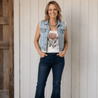 Woman wearing a denim vest, floral tank top, and jeans standing against a wooden door.