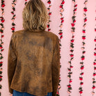 A person viewed from behind wearing a brown distressed leather jacket and blue jeans, standing against a pink backdrop decorated with vertical strands of red and pink artificial flowers.