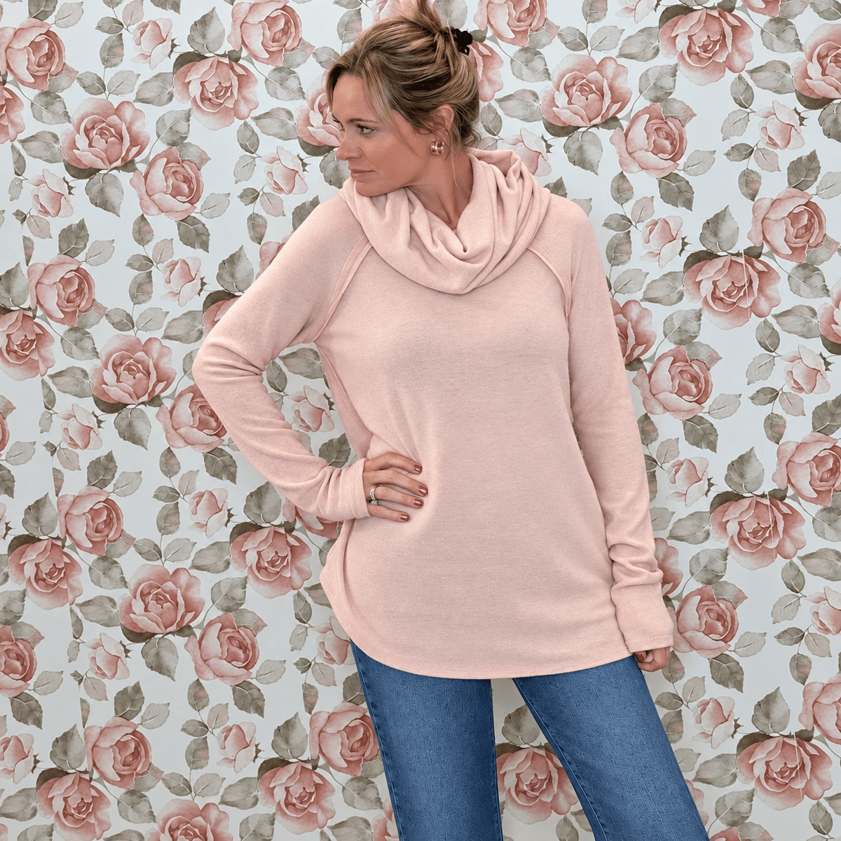 Crystal wearing a smoke pink brushed knit cowl neck top with raglan long sleeves and jeans, in front of rose wallpaper