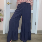 A woman posing in a pink and white vertically striped long-sleeve button-up shirt and navy blue wide-leg tiered pants, standing in front of a white door inside what appears to be a boutique.