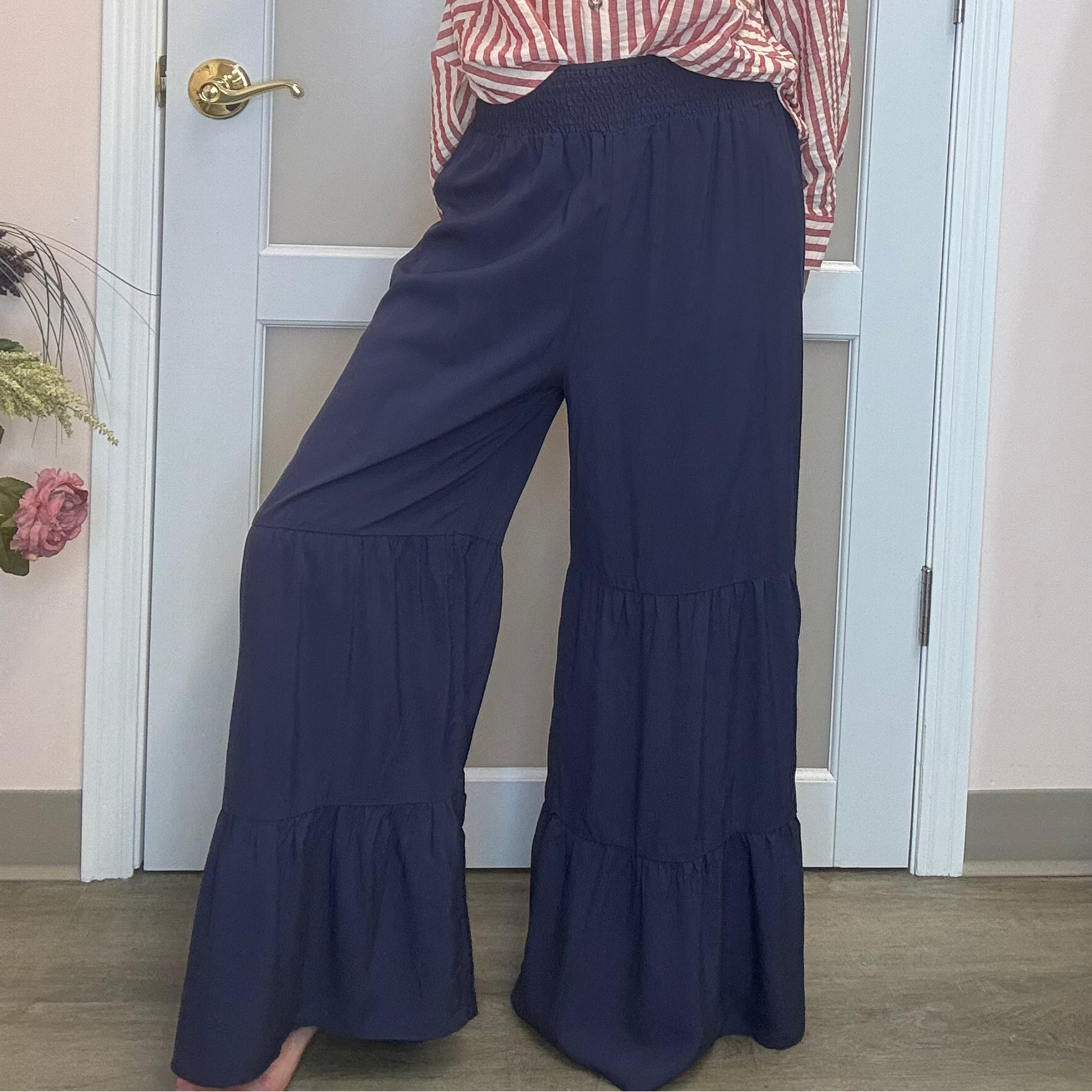 A woman posing in a pink and white vertically striped long-sleeve button-up shirt and navy blue wide-leg tiered pants, standing in front of a white door inside what appears to be a boutique.