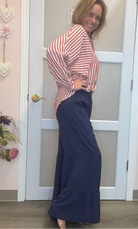 A woman posing in a pink and white striped long-sleeve top with navy blue tiered maxi pants, standing in front of a white door in a boutique setting.