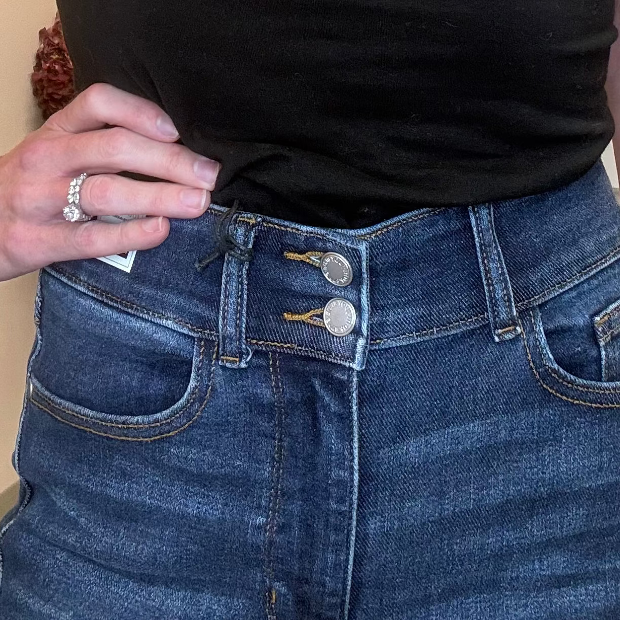 Close-up of a person wearing dark blue denim jeans with a button fly and silver buttons, paired with a black top. The person's hand with a ring is resting on the waistband.