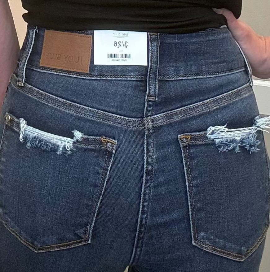 A woman viewed from behind wearing a black short-sleeve t-shirt and high-waisted blue jeans with distressed back pockets, standing indoors.