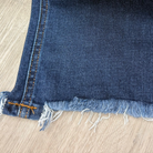 A close-up of a dark blue denim fabric with frayed edges and orange contrast stitching, laid flat on a light wood floor.