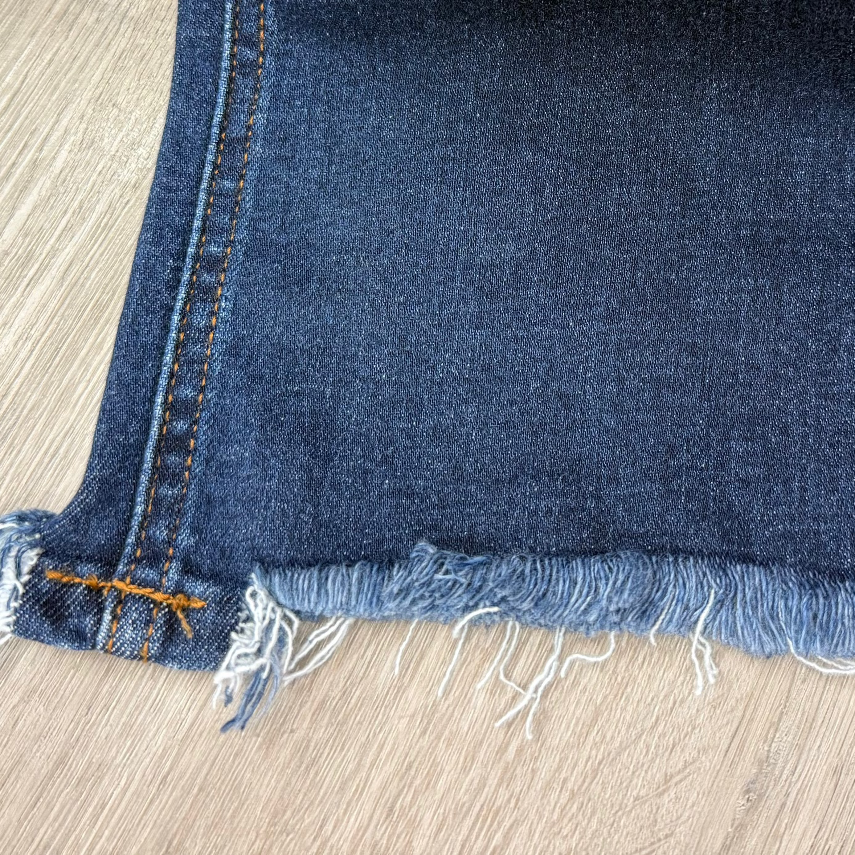 A close-up of a dark blue denim fabric with frayed edges and orange contrast stitching, laid flat on a light wood floor.