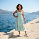 Woman in a light blue dress standing by a waterfront with mountains in the background