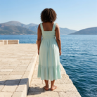 Woman in a light green dress standing on a dock by the water with mountains in the background