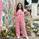Woman in pink floral overalls standing in front of a colorful mural with flowers around.