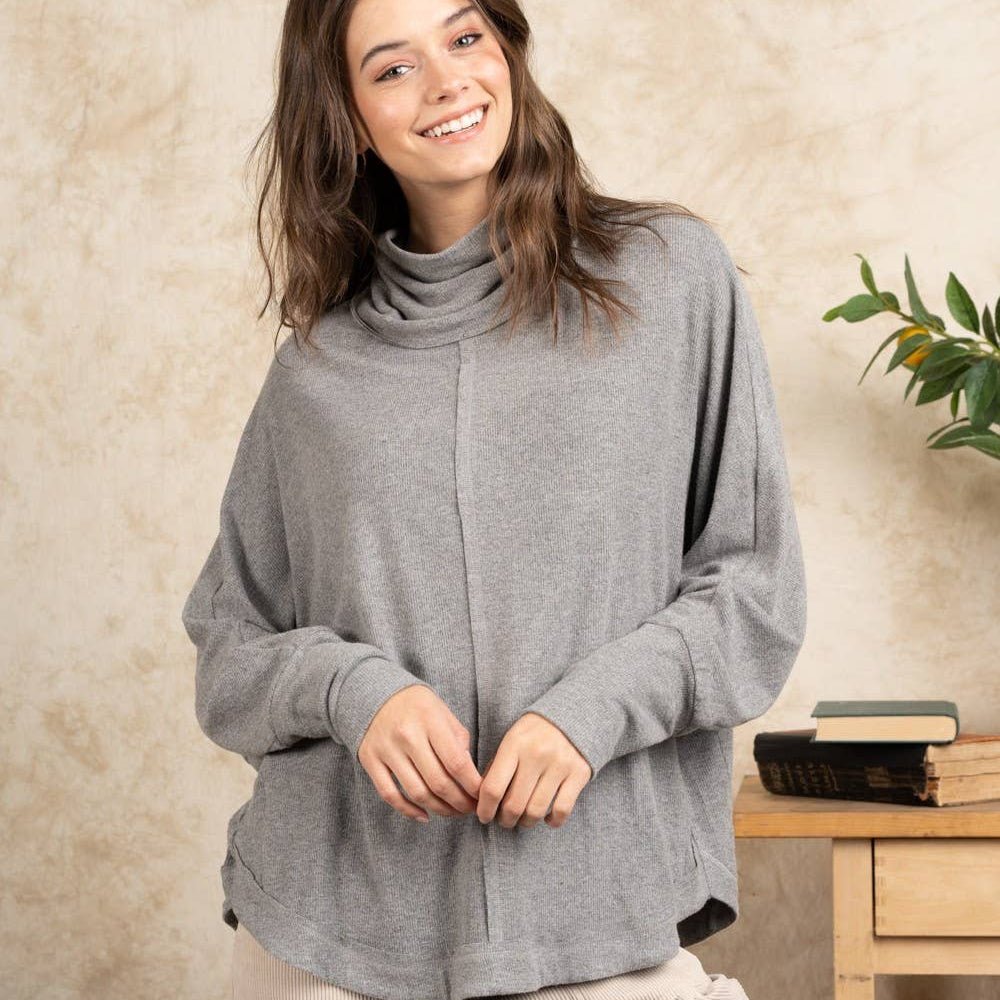 A woman wearing a heather gray cowl-neck knit sweater and beige cargo pants, standing in front of a textured beige wall with a small wooden side table and green plant in the background.
