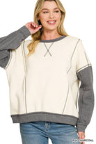 Woman wearing an oversized cream fleece pullover with heather charcoal sleeves, exposed seams and V-stitch neckline, styled with light wash jeans