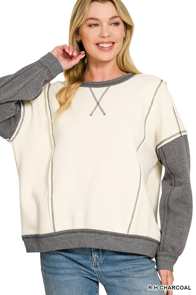 Woman wearing an oversized cream fleece pullover with heather charcoal sleeves, exposed seams and V-stitch neckline, styled with light wash jeans