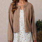 Woman wearing a brown cardigan over a white floral dress against a beige wall.