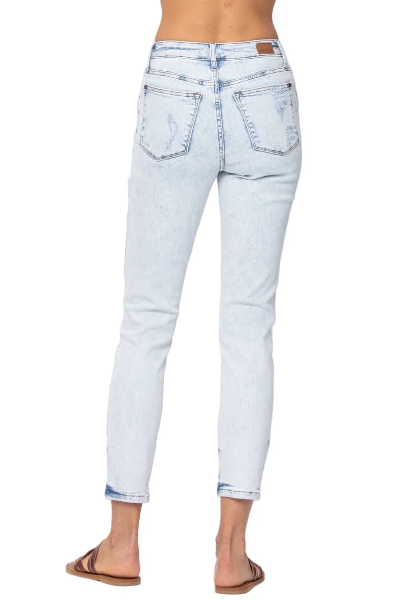 Rear view of a person wearing light-wash blue cropped denim jeans with rolled cuffs and flat brown slide sandals, against a plain light gray studio background.