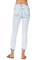Rear view of a person wearing light-wash blue cropped denim jeans with rolled cuffs and flat brown slide sandals, against a plain light gray studio background.