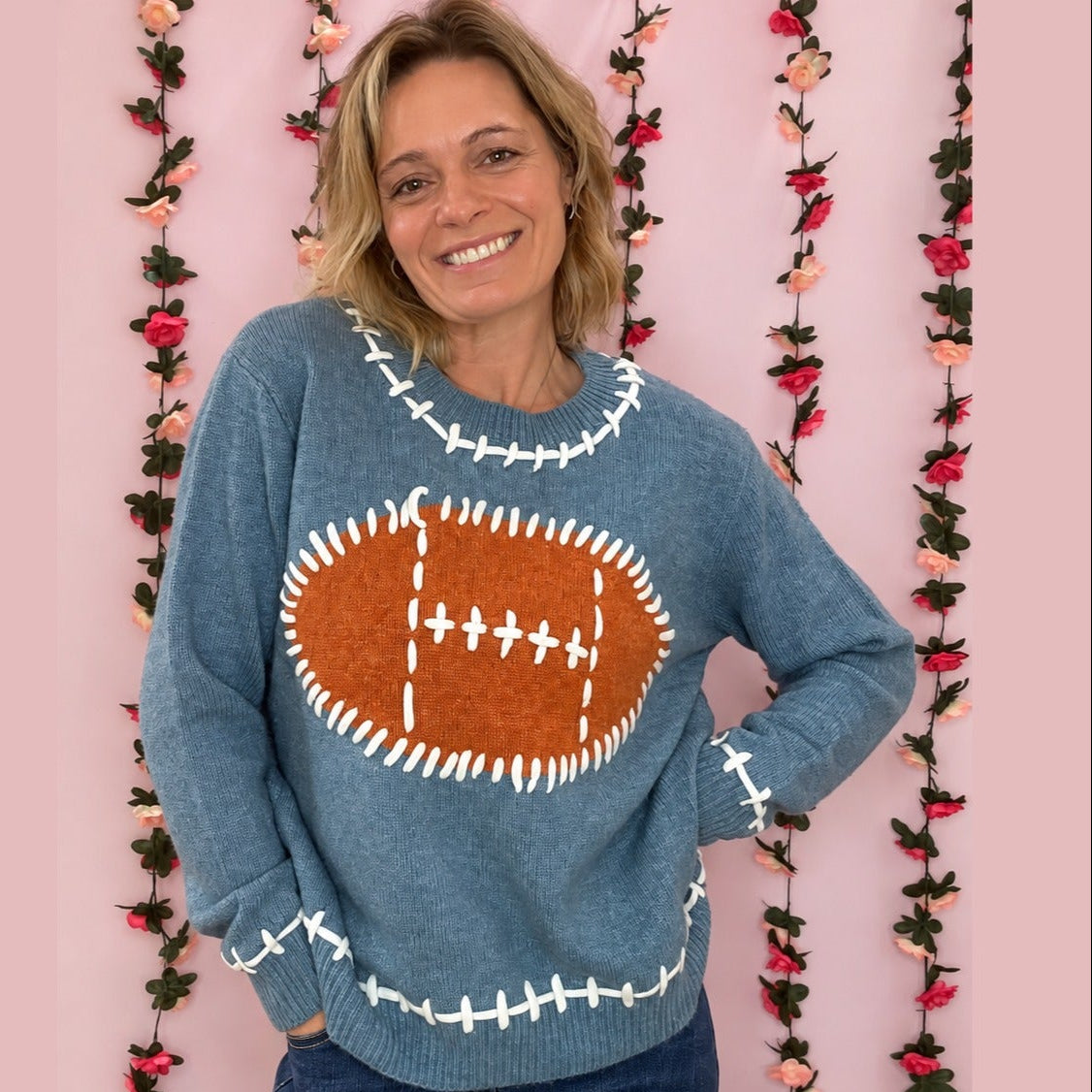 Woman wearing a dusty blue football knit sweater with a brown football graphic and white stitch details