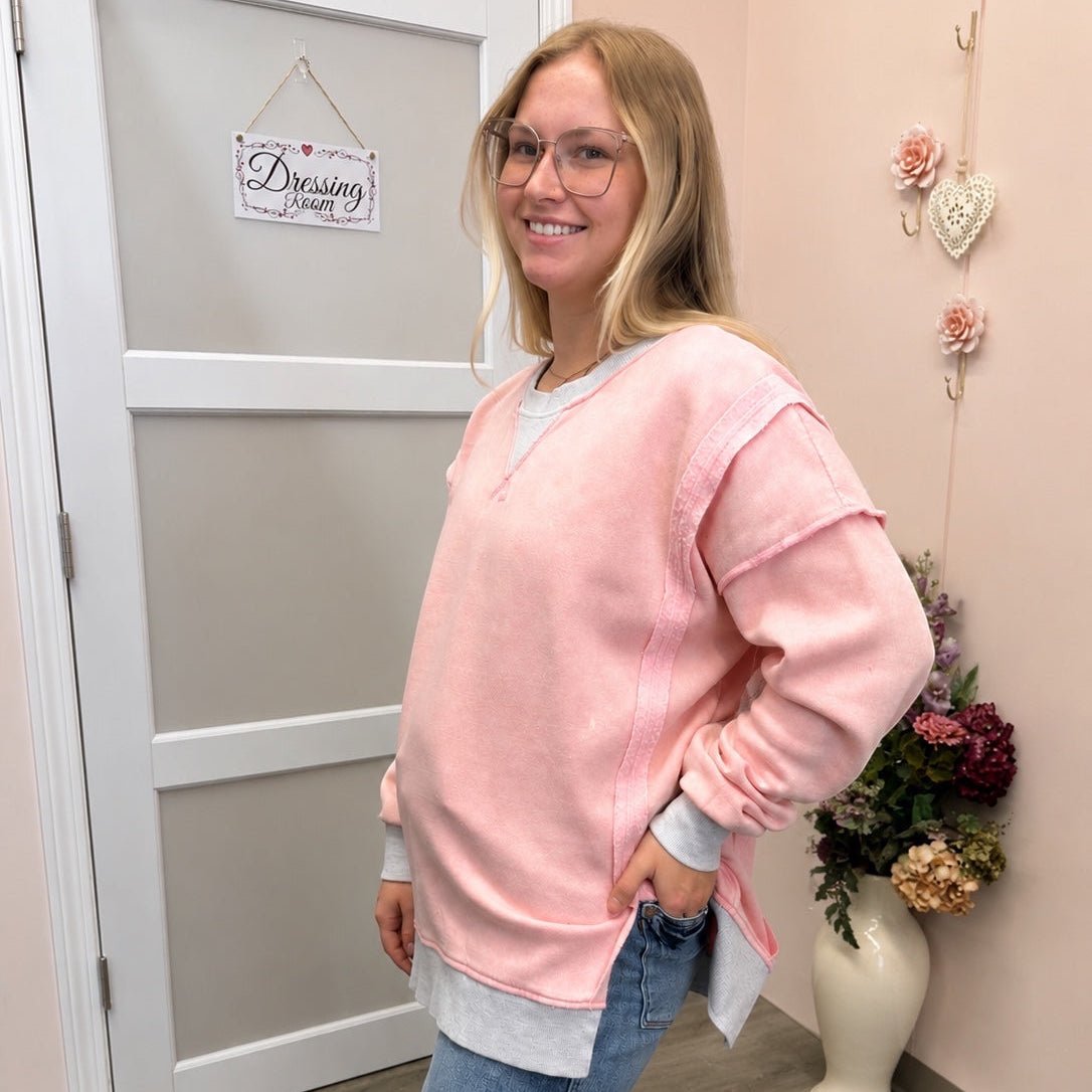 A woman modeling a light pink long-sleeve crewneck sweatshirt layered over a white undershirt, paired with blue jeans, standing in an indoor room with a beige wall, white door, and a vase of flowers.