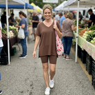 Woman in brown t-shirt and brown biker shorts carrying a pink cherry bag.  she is walking thru a farmers market.