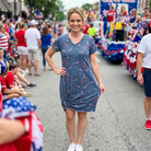 Woman in a blue dress with red and white pattern standing in a parade with people and flags in the background.