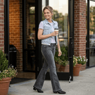 Woman walking outside a building holding a coffee cup. Wearing Black jeans and purple t-shirt with a denim vest.