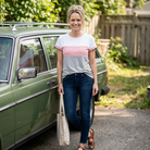 Woman standing next to a green car holding a beige tote bag, wearing a colorful striped shirt and blue jeans.