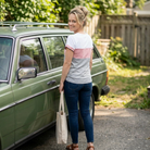 Woman standing next to a green car holding a tote bag, with trees and a wooden fence in the background.