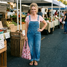 Woman standing at an outdoor farmers market holding a pink bag wearing overalls