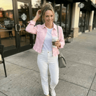 Woman in pink jacket and white jeans holding a coffee cup on a sidewalk.