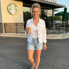 Woman holding a Starbucks drink in front of a Starbucks store wearing a white denim jacket and white shirt with light denim shorts.