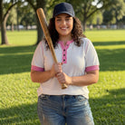 Woman holding a baseball bat in a park wearing a white shirt trimmed in pink.