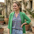 Woman in a green jacket and floral shirt standing in an outdoor setting with stone walls and potted plants.