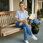 Woman sitting on a wooden bench holding a drink on a porch with flowers and plants.