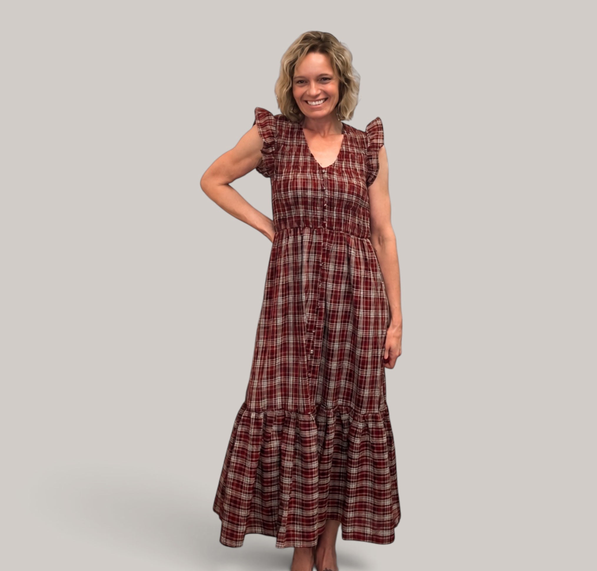 A woman modeling a long-sleeved, tiered maxi dress with a red and brown plaid pattern, set against a solid light gray background.